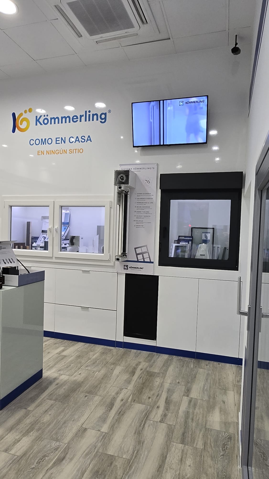 Exposición de ventanas y sistemas de cerramientos Kömmerling en Alicante. La imagen muestra un área de exhibición moderna con ejemplos de ventanas corredizas y abatibles, junto con un modelo de sistema de persianas integrado (Sistema Kömmerling76). En la pared, el eslogan 'Como en casa, en ningún sitio' resalta la marca, acompañado por una pantalla que muestra productos en funcionamiento. Los muebles blancos y el diseño limpio enfatizan el enfoque en tecnología y estética